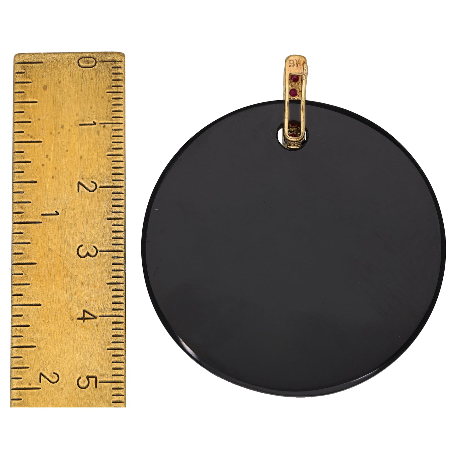 Round black onyx pendant with gold swirl and ruby red stone set bail, shown with ruler